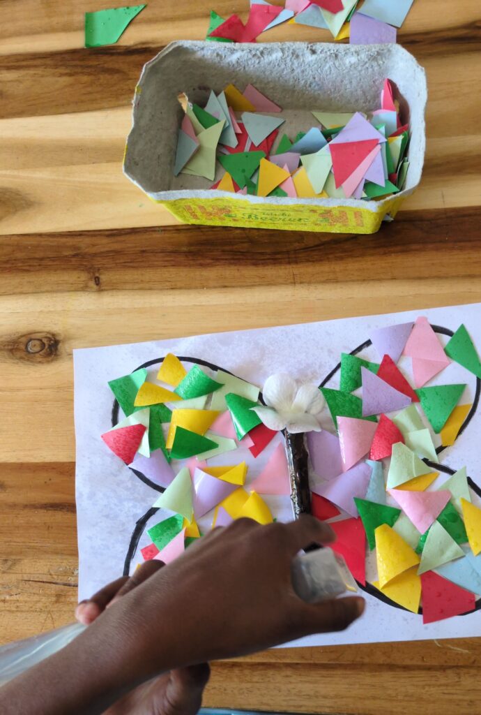 Preschooler spraying water on colored bleeding tissue paper squares inside a hand-drawn butterfly outline for a summer sensory craft.