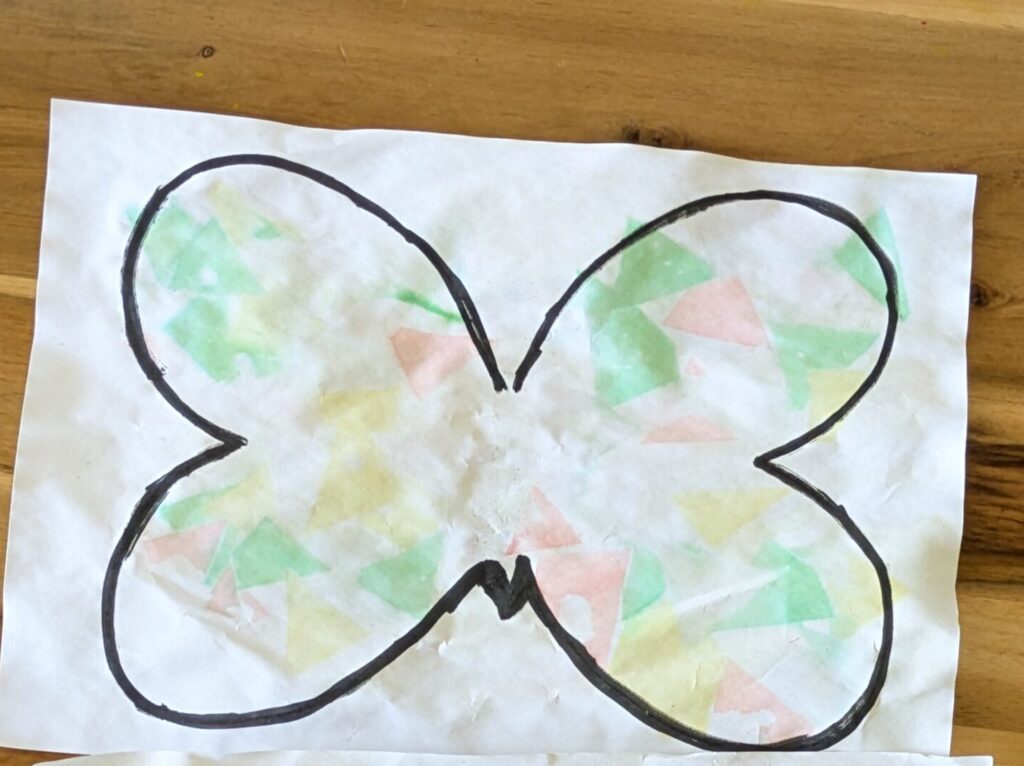 Preschooler spraying water on colored bleeding tissue paper squares inside a hand-drawn butterfly outline for a summer sensory craft.