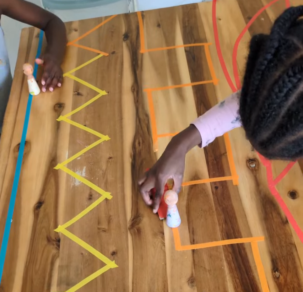 Small wooden toy figures being moved along a zigzag painter's tape track on the floor for a toddler coordination and concentration activity.