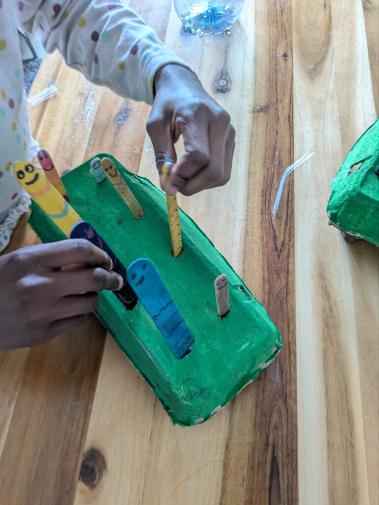 A recycled cardboard egg carton painted brown and green with face-drawn popsicle sticks inserted as worms for a toddler fine motor activity.