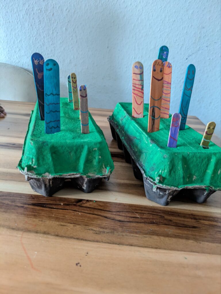 A recycled cardboard egg carton painted brown and green with face-drawn popsicle sticks inserted as worms for a toddler fine motor activity.